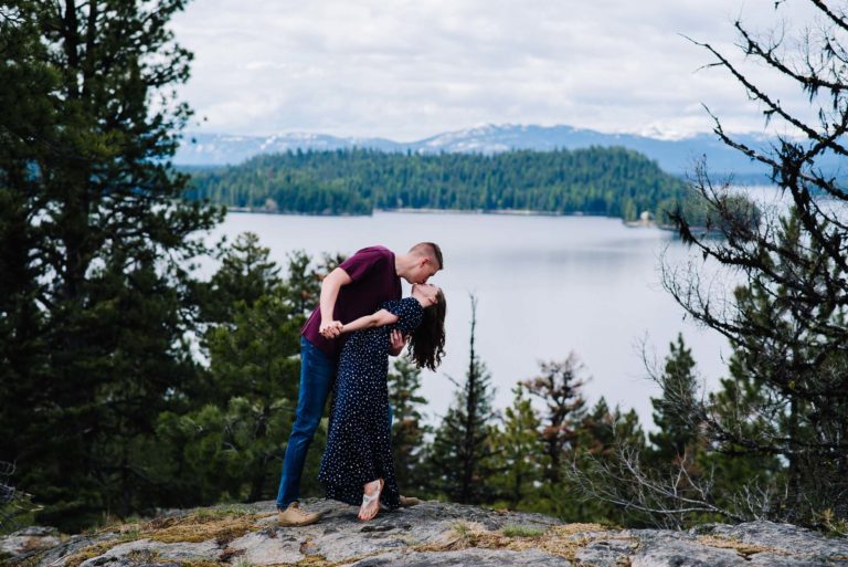 A couple kisses with Payette Lake in the background. There are snow caped mountains and pin trees on the lake. This location is one of the best engagement photo locations in Boise. 