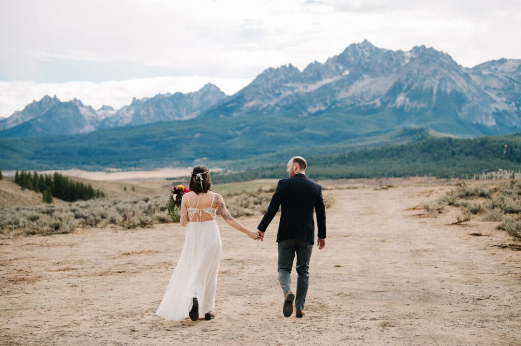 A couple walks away from the camera holding hands in Idaho. There is an expansive mountain landscape in front of them. The groom is wearing a blue blazer and the bride is wearing an open back dress.