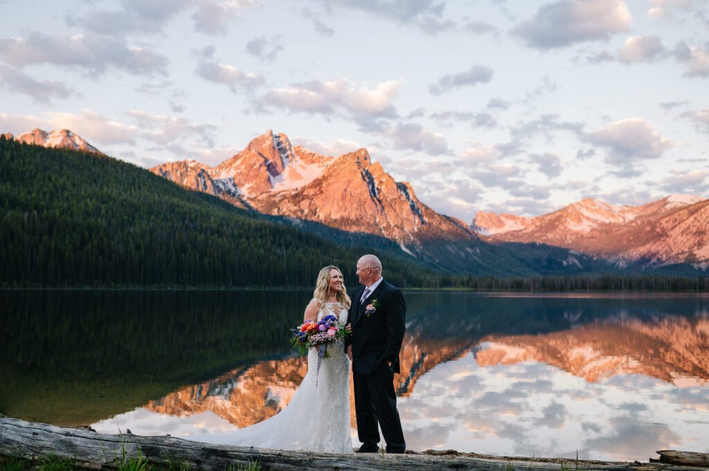 A couple stands together during their first look at sunrise on Stanley Lake. The rising sun is casting an alpenglow on the mountains behind the lake. The orange and pink mountains are reflecting on the lake.