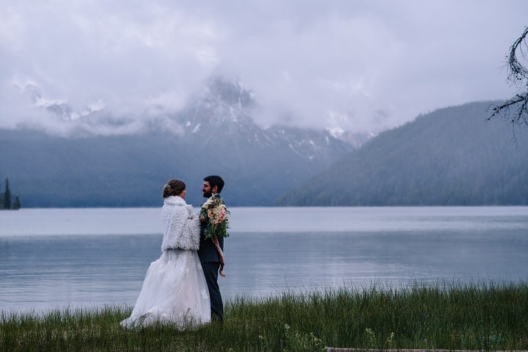 Wedding would stands with sawtooth mountains behind them during their Bride and groom embrace with snow falling around them at their Redfish Lake Lodge Wedding in Stanley, Idaho.