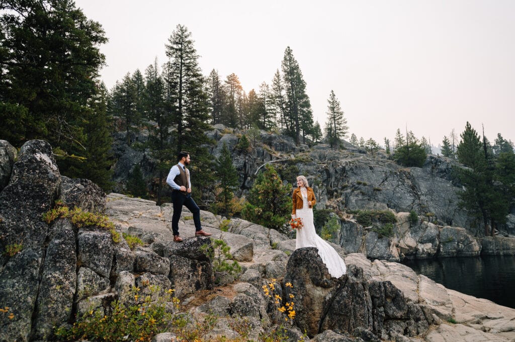 A bride and groom stand apart on two rocks. The bride is wearing an orange leather jacket. This couple eloped in McCall, Idaho.