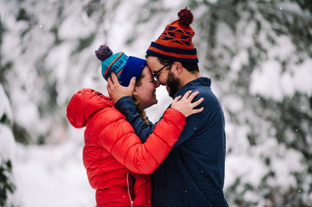 Couple smiles with foreheads touching in a snowy forest.