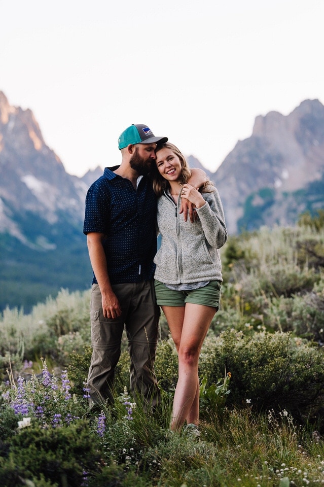 A couple stands together on a ridge with mountains behind them. This couple wore nice looking hiking cloths for their engagement session in the mountains of Stanley, Idaho.