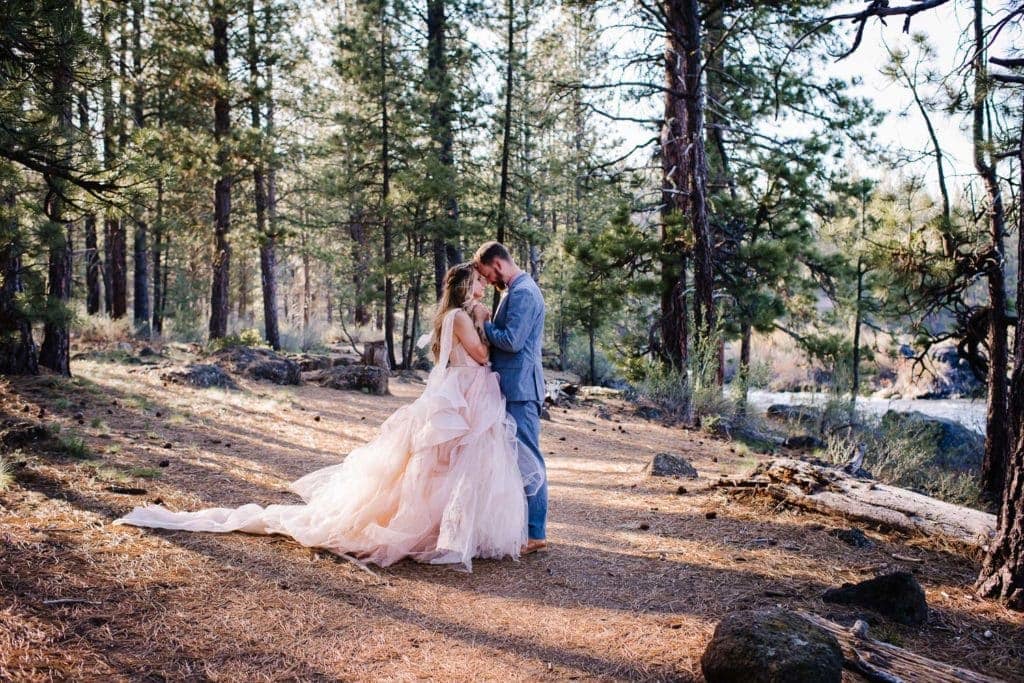 Bride and groom stand touching foreheads and holding hands. Couple is standing in a pine tree forest. Bride is wearing a pink dress and groom is wearing a light blue suit.