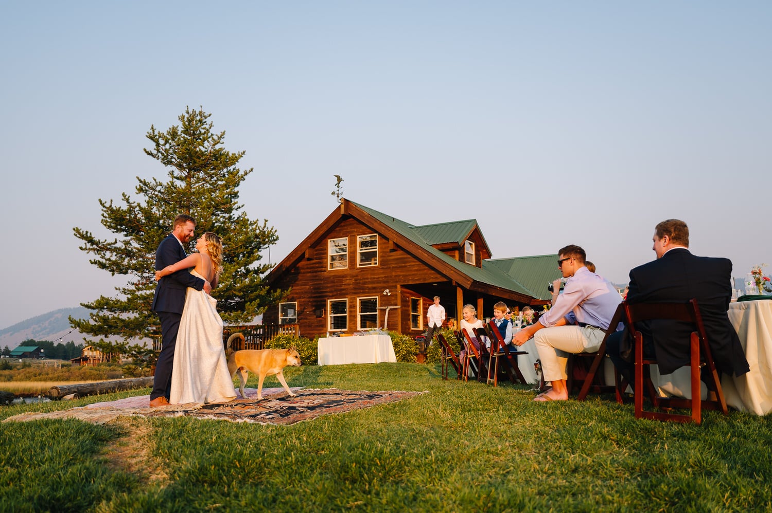 A wedding couple shares their first dance in front of a Airbnb cabin in Stanley, Idaho. There are wedding guests sitting along a table watching the couple dance.