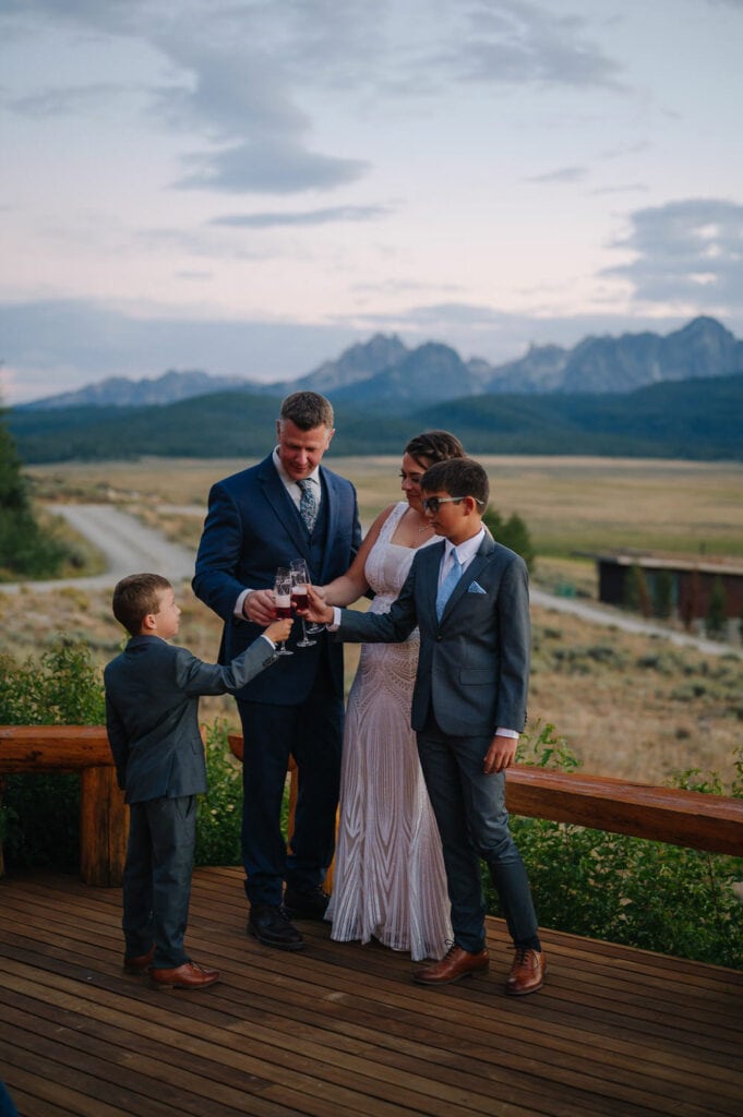 A family stands together on the porch of their Airbnb wedding venue in Idaho. They are enjoying a celebratory drink at sunset. There are mountains behind them.