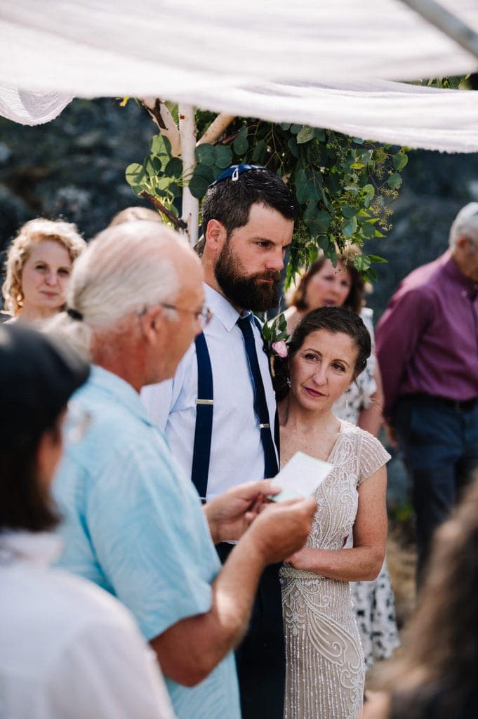 Father of the groom reads the Seven Blessings during a Jewish-Catholic wedding ceremony. Wedding couple is standing under a chuppah and surrounded by their family.