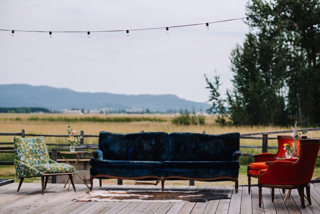 A blue velvet antique couch and orange velvet antique chairs are used to decorated a reception space at the Historic Roseberry Barn in McCall, Idaho. There are mountains and trees in the back ground. 
