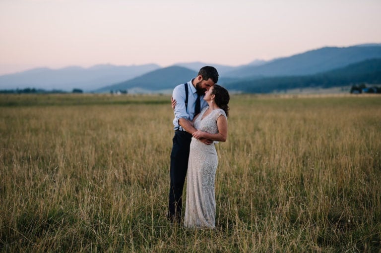 Bride and groom stand together in a mountain meadow after their McCal, Idaho wedding. There is tall grass and mountains surrounding them.