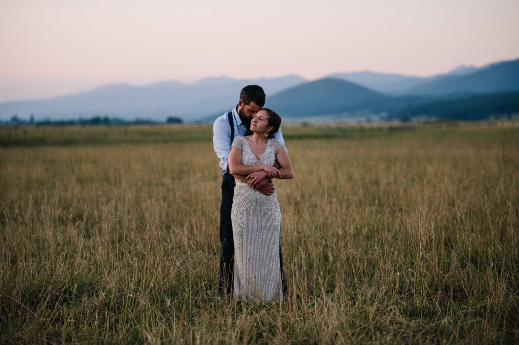 Bride and groom stand in a grassy field at sunset with mountains behind them. This couple planned a reception at the Historic Roseberry Barn in McCall, Idaho.