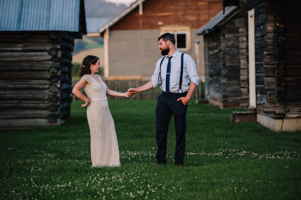 Bride and groom stand holding hands along old log cabins at the Historic Roseberry Barn wedding venue in McCall, Idaho.