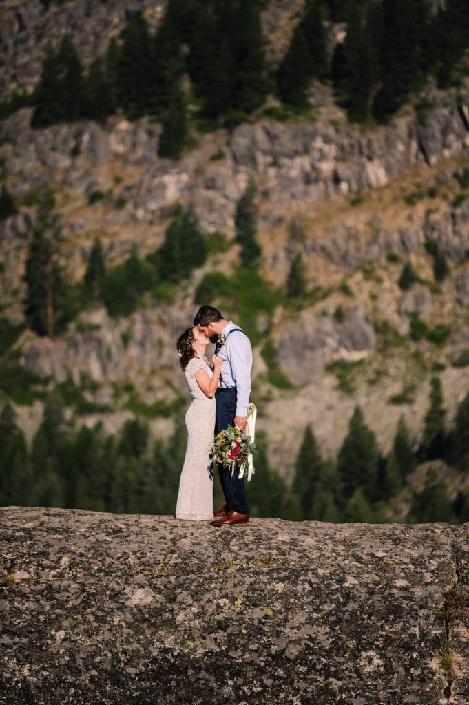 Bride kisses her groom in the early morning sunlight during their adventure session before their wedding ceremony. Couple is standing on a rock with trees and mountains behind them.