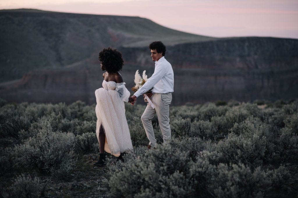Elopement couple walks across a sagebrush outcropping on the Snake River near Boise, Idaho. Groom is holding a bouquet of flowers and bride is leading groom. Boise is one of the best places to elope in Idaho.