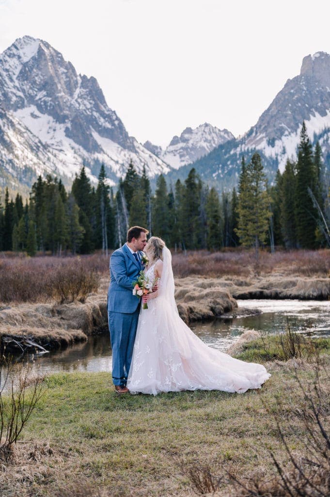 Couple kisses after their elopement ceremony in Stanley, Idaho. Couple is standing in a meadow with snow capped mountains and a creek behind them. 