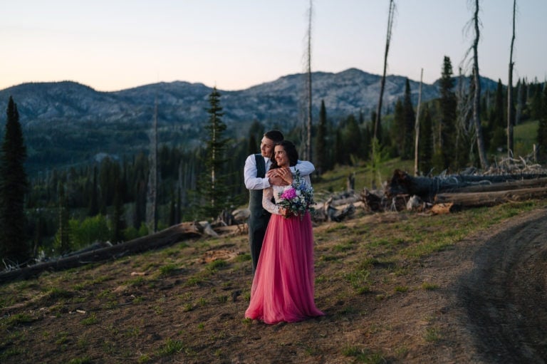 Couple stands together with the setting sun on their faces during their McCall elopement. There are mountains behind them. Bride is wearing a pink skirt and the groom is wearing a dark green suit.