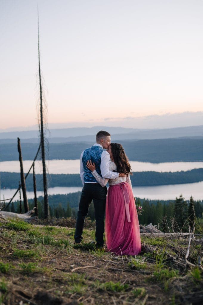 Couple stands with their backs to the camera. Groom is kissing the bride on the forehead. There are mountains and a lake behind the couple. 