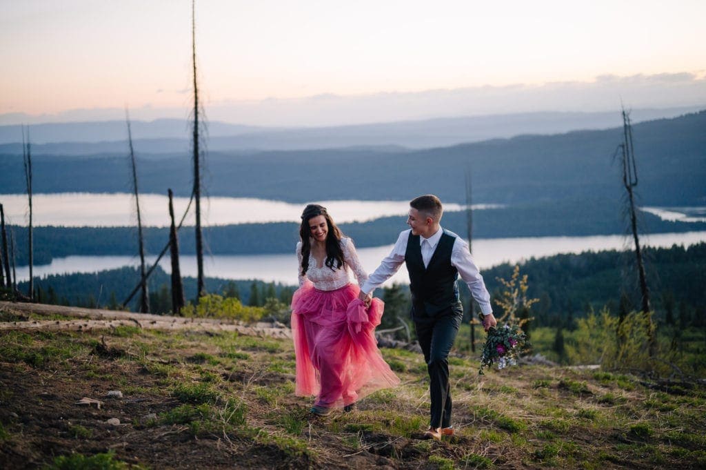 Couple walks up a hillside holding hands in the late evening light. Bride is wearing a pink tulle skirt and bodysuit from Sweet Caroline Styles. Groom is wearing a green suit from Men’s Wearhouse and holding a bouquet.