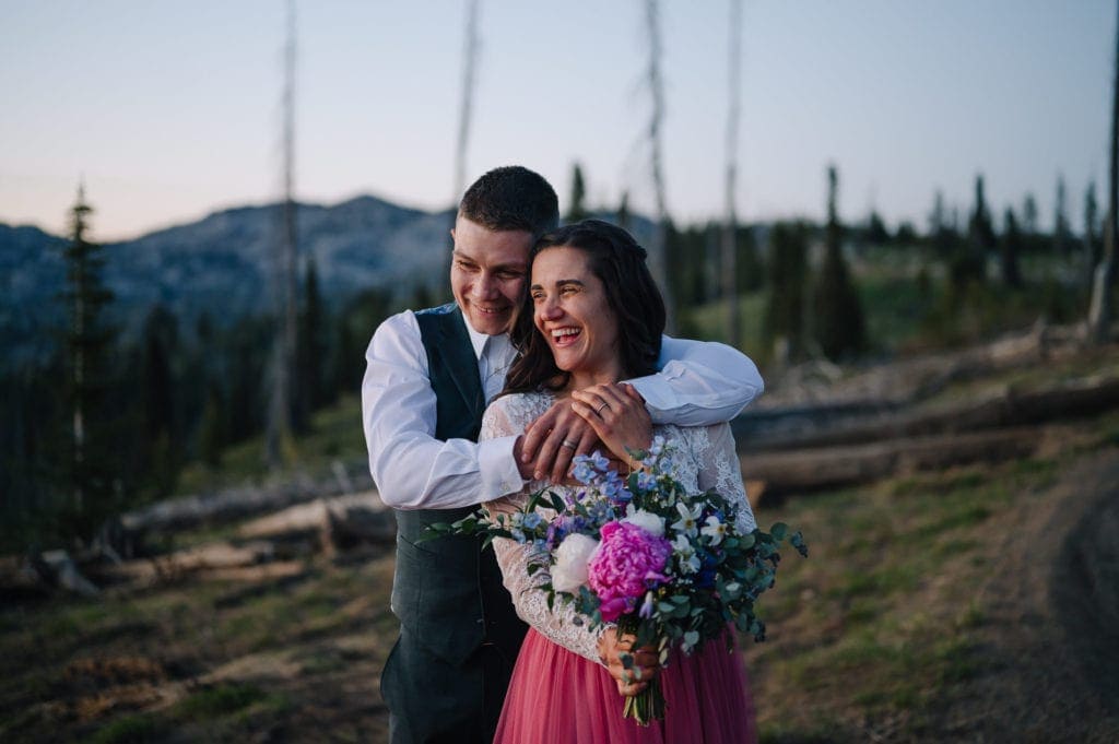 Groom embraces bride. Couple is laughing and the bride is holding a bouquet of flowers. Couple is standing in the mountains above McCall, Idaho.