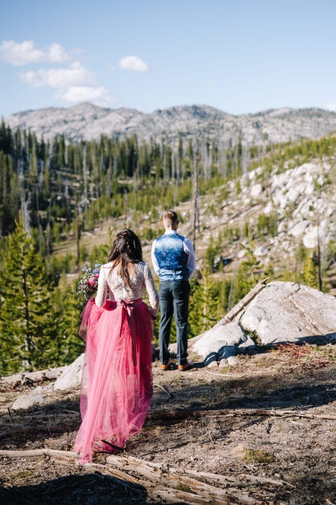 Bride walks up behind the groom during their first look. This couples first look was done at the top of the mountain before their elopement ceremony. Bride is wearing a pink skirt and white bodysuit. Groom is in a green suit. There are mountains and trees behind the couple.