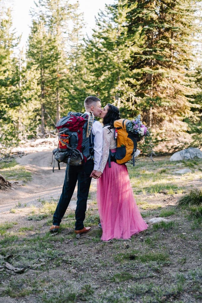 Bride and groom kisses during their elopement hike. Couple is dressed in wedding attire and wearing backpacks. Couple is standing in a forest of Pine Trees and the sunlight is coming through the trees behind them.