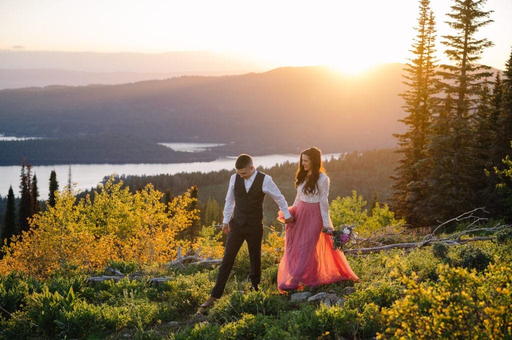 Couple walks together along a mountain ridge in the setting sun. There is a lake and mountains in the distance. Bride is wearing a pink tulle skirt and bodysuit from Sweet Caroline Styles and the groom is wearing a green suit from Men’s Wearhouse. Bride is holding a bouquet from a McCall, Idaho florist.