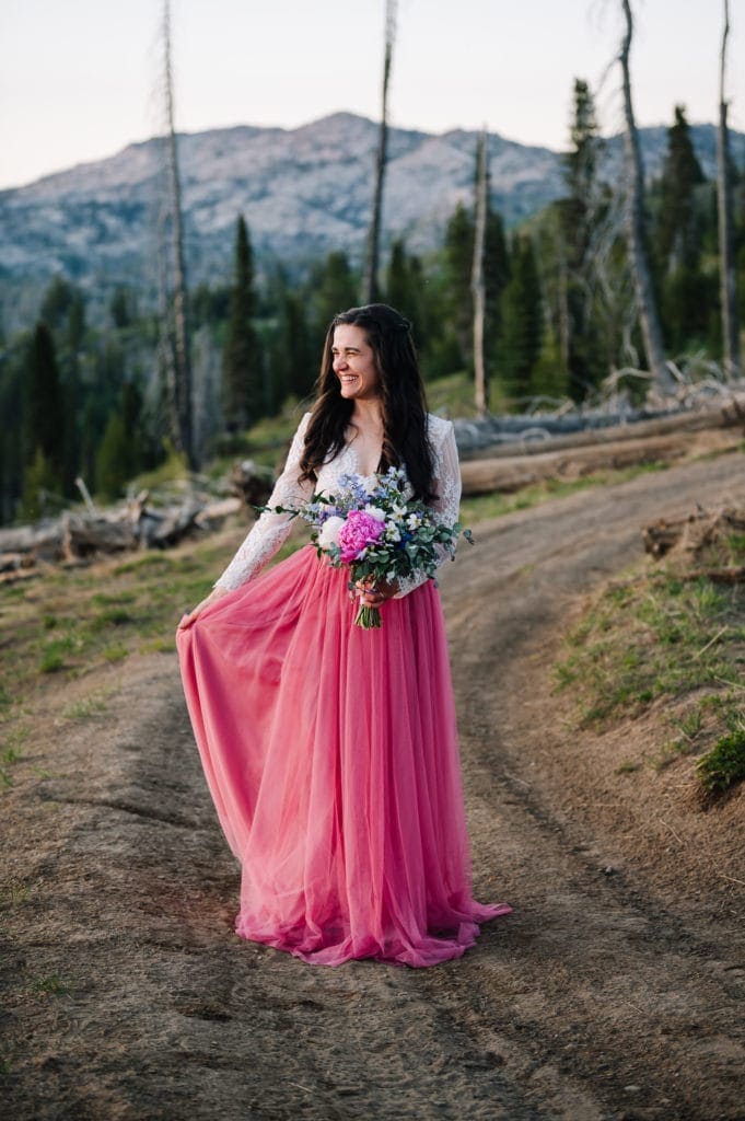 Bride stands holding the side of her dress on one hand and a bouquet of flowers in her other hand. Bride is wearing a pink tulle skirt and bodysuit from Sweet Caroline Styles. She is looking to the side of the photo and smiling. 