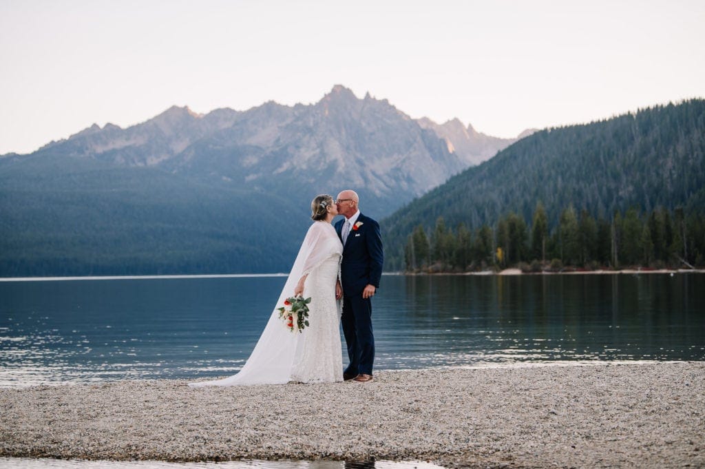 A couple kisses during their Idaho elopement at Redfish Lake Lodge. The sun is setting on the lake behind the couple. Groom is wearing a blue suit and bride is wearing a white dress and bridal cape.