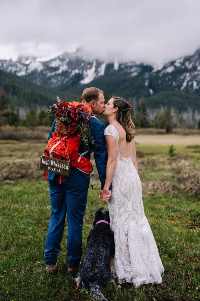 A couple kisses during their Idaho elopement in Stanley. The groom is wearing a backpack with flowers and a sign that reads 'just married'. The couples dog is standing between them. There are mountains covered in fog and a meadow behind the couple.