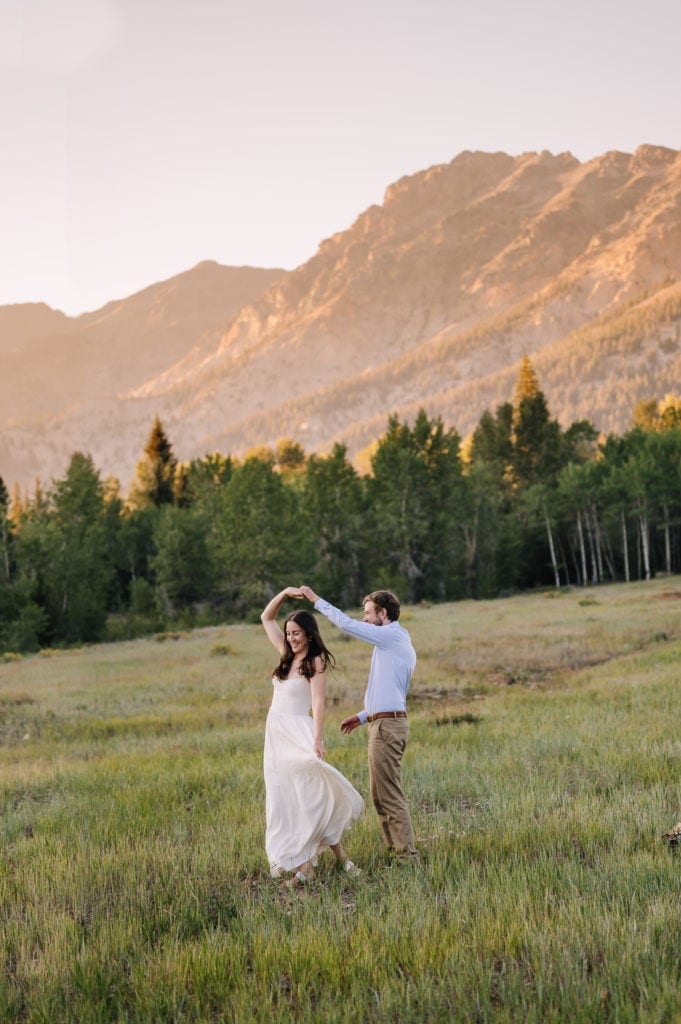 A couple dances in a mountain meadow during their Idaho elopement in Sun Valley. The evening sun is setting on the mountains behind the couple. The groom is wearing a blue shirt and khaki pants. The bride is wearing a cream dress from Reclamation.