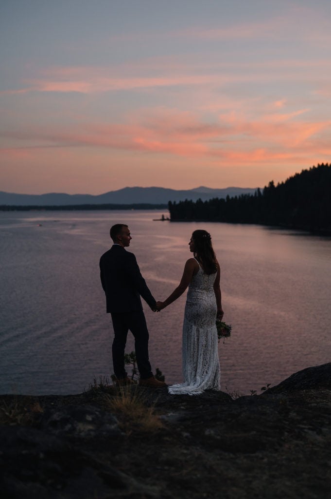 A couple stands at sunset holding hands during their Idaho elopement in McCall. The couple is s silhouetted against Payette Lake. The sunset is pink behind the couple.