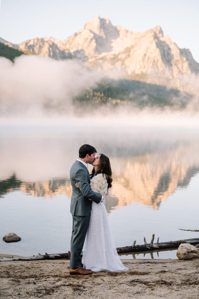 An eloping couple exchanges their first kiss on an alpine lake in Stanley, Idaho. They are standing on a white sand beach and the mountains are reflecting in the water hind them.