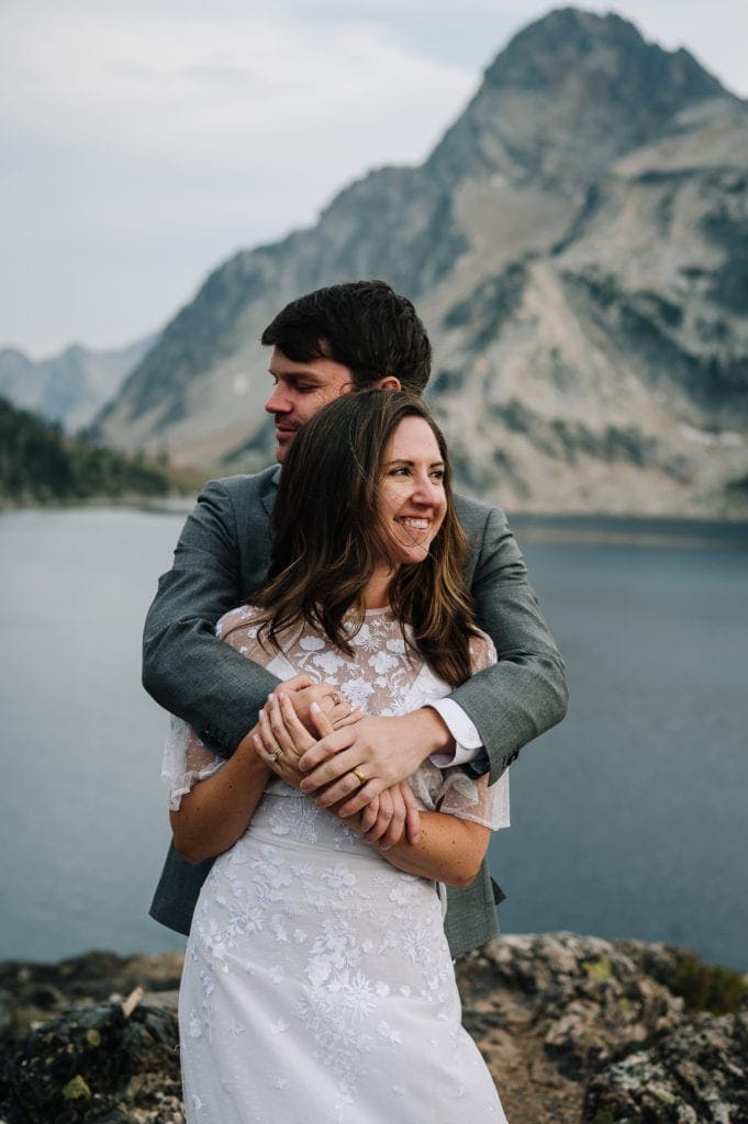 Groom hugs bride from behind. Couple is looking away from the camera and there is a mountain and alpine lake behind them.