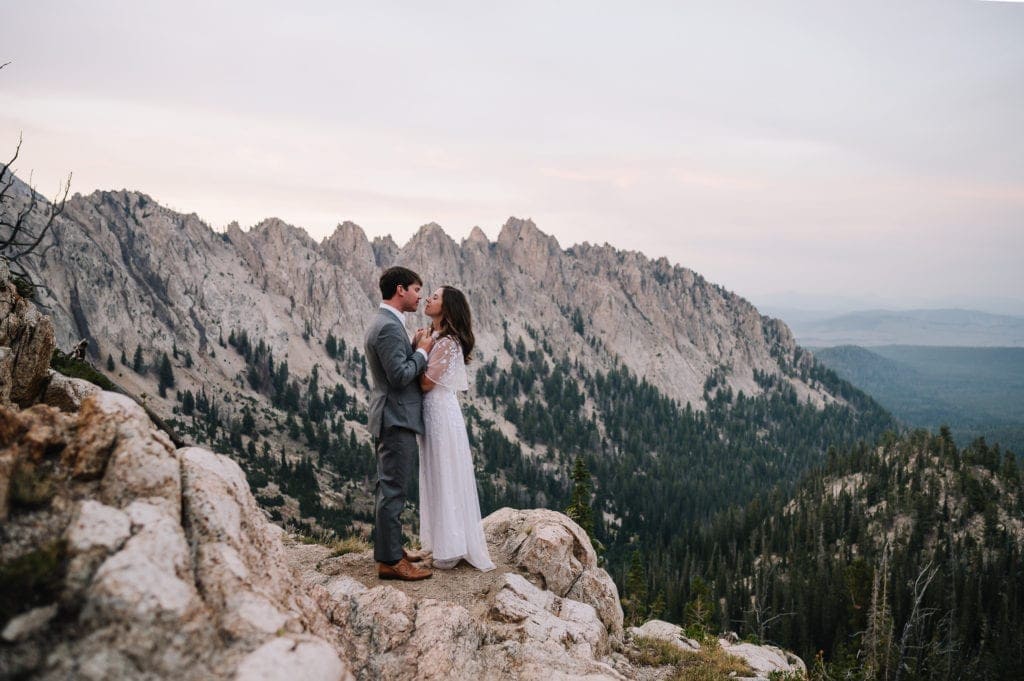 A couple stands on the edge of a cliff with the Sawtooth Mountains behind them. This photo was taken at sunset during their Idaho wedding adventure.