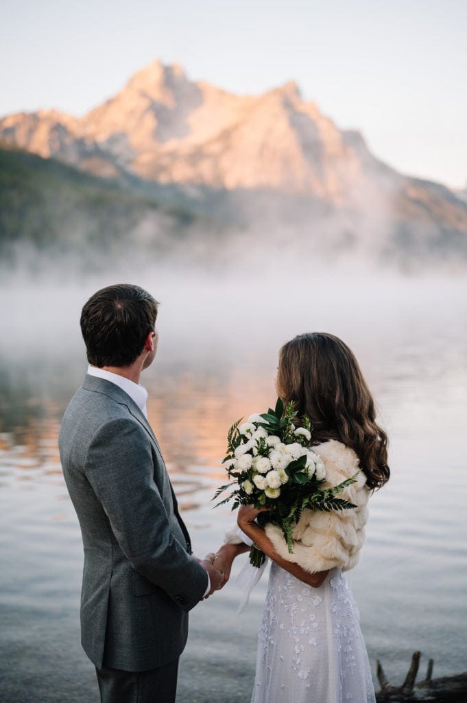 Bride and groom stand holding hands and looking at the mountains around them during their elopement ceremony. Their backs are to the camera. Bride is holding a bouquet of white Mums.