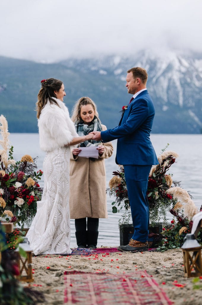 An eloping couple smiles at each other during their Redfish Lake wedding ceremony. Couple is standing on a rug and there are large pampas grass and red Ranunculus flower arrangements on either side of the couple. Cloudy mountains and a lake are visible behind the couple. Groom is wearing a blue suite with rain boots. Bride is wearing a white, faux furry quarter length sleeve jacket and a white lace dress.