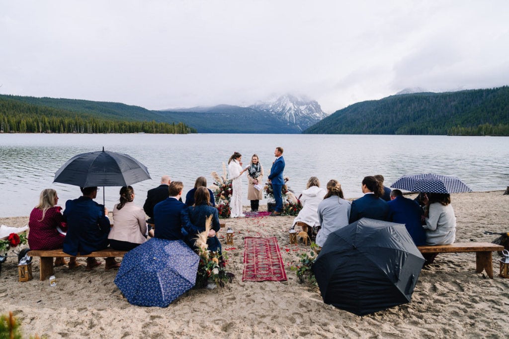 A couple exchanges vows during their Redfish Lake wedding ceremony. The couple is standing on a beach with their wedding guests. Guests are seated on wooden benches and covers with umbrellas. Cloudy mountains and pine trees are visible behind the couple.