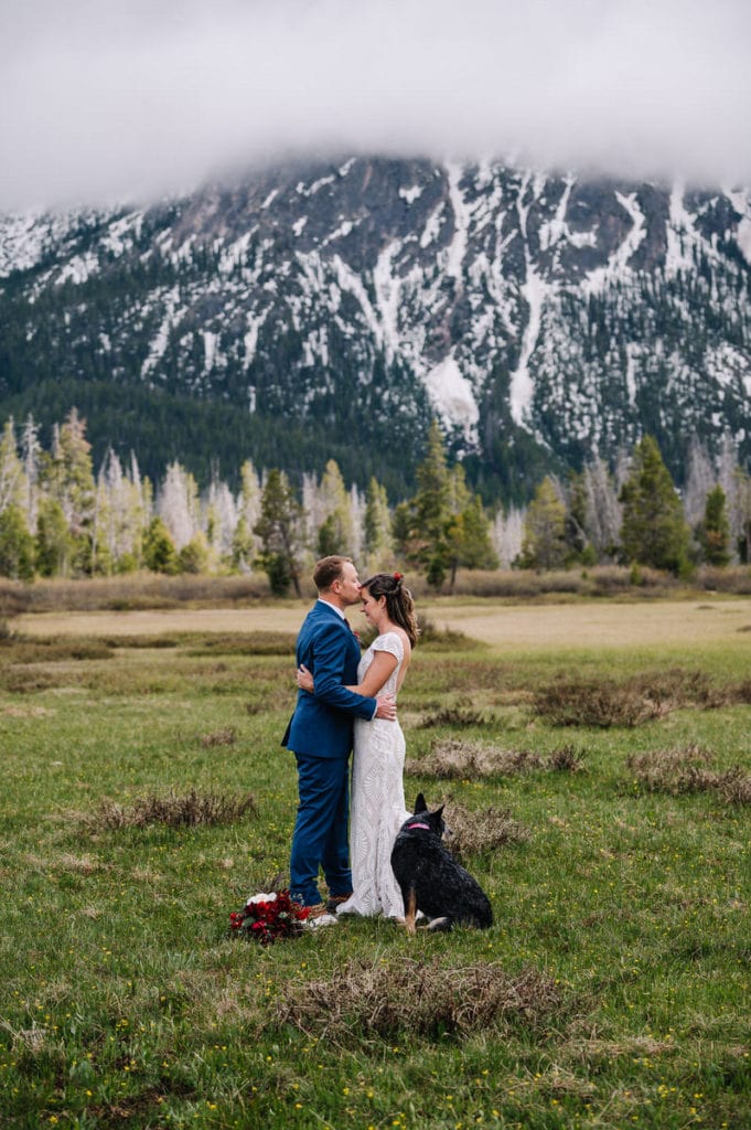 Groom kisses brides forehead during their elopement in Stanley, Idaho. Couple is standing in a green mountain meadow with cloudy mountains behind them. There is snow on the mountain and the couples dog of honor is sitting at their feet.