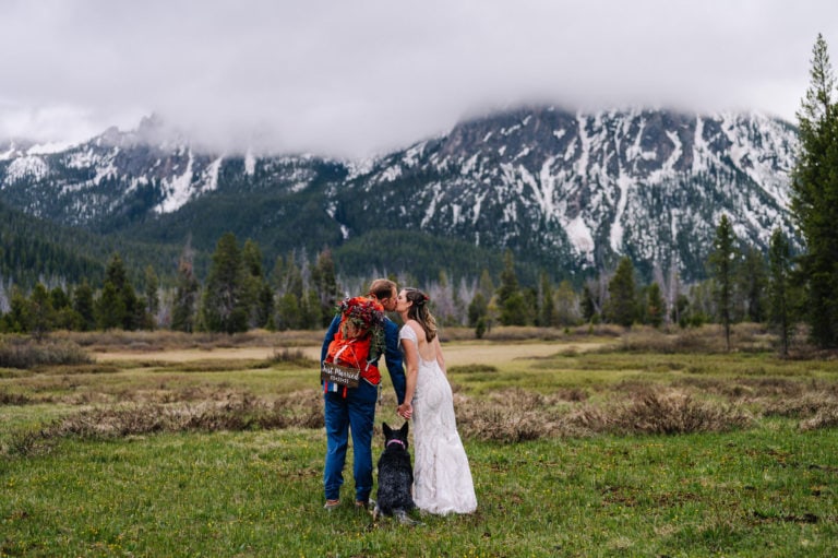 A couples kisses with their dog standing close during there Redfish Lake Wedding. Couple is standing in a green mountain meadow with cloudy mountains surrounding them. Groom is wearing a blue suit and an orange backpack. The backpack holds a the brides bouquet and a sign that says 'just married'. Bride is wearing a white lacy, open backed dress.