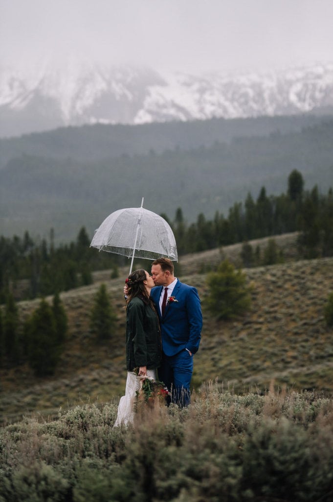 Couple kisses under a clear umbrella during their Stanley, Idaho elopement. Couple is standing on a hillside covered with short sagebrush. There are cloudy mountains covered with snow behind the couple.