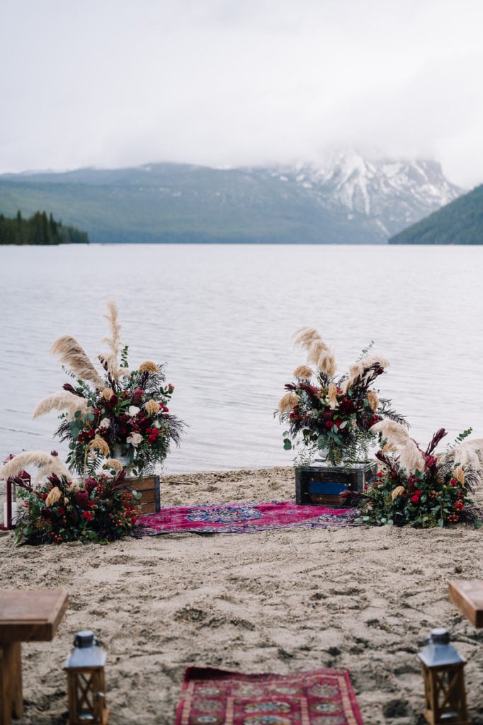Photo of a ceremony arch setup for a Redfish Lake wedding. There are two large floral arrangement on either side of the aisle that include pampas grass, Seeded Eucalyptus greenery and red Ranunculus flowers.
