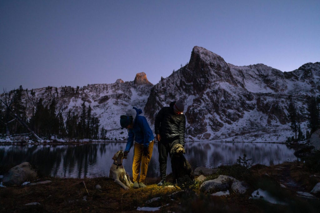 Couple stands looking down at their dogs during their twilight elopement wedding in Idaho. Bride and groom are wearing headlamps and shining light on their dogs. Light from the rising moon can be seen on the mountains and lake behind the couple. 