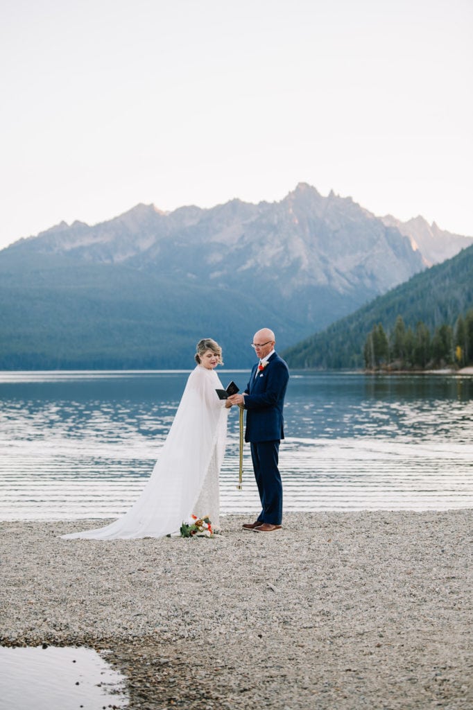 A couple performs a handfasting ceremony during their Idaho elopement package. The couple is standing on a beach at Redfish Lake. There are mountains behind them.