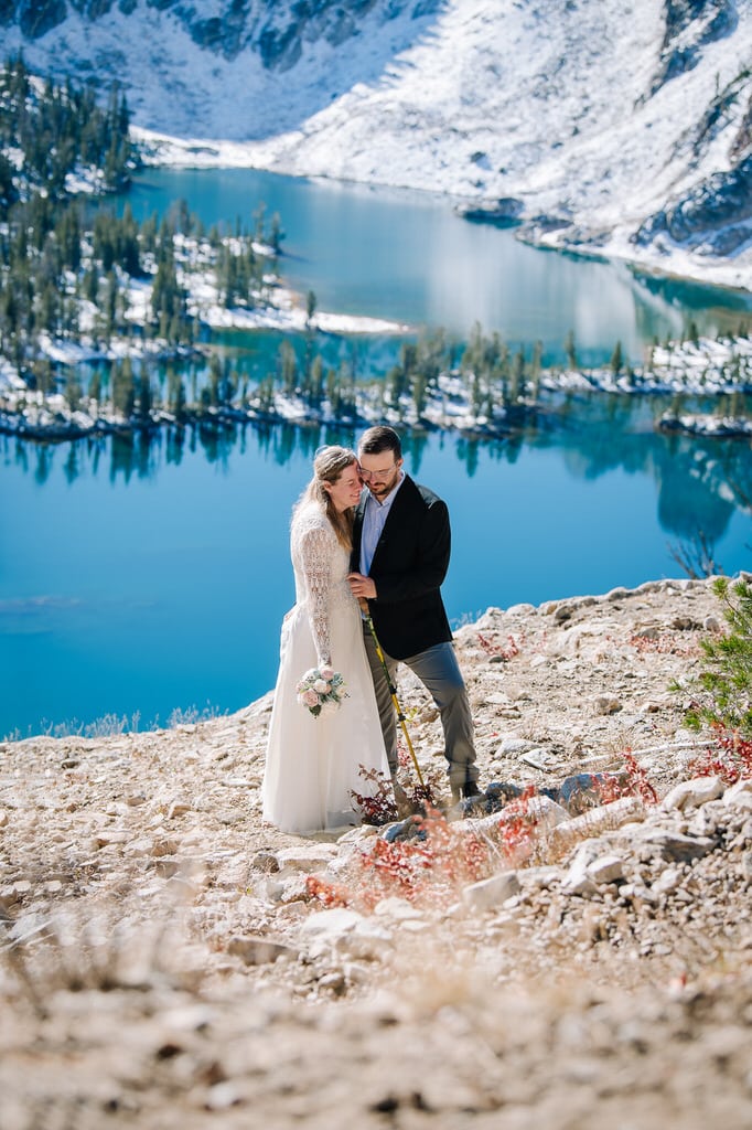 A couple stands together with an aqua blue lake behind them during their Idaho backpacking elopement.
