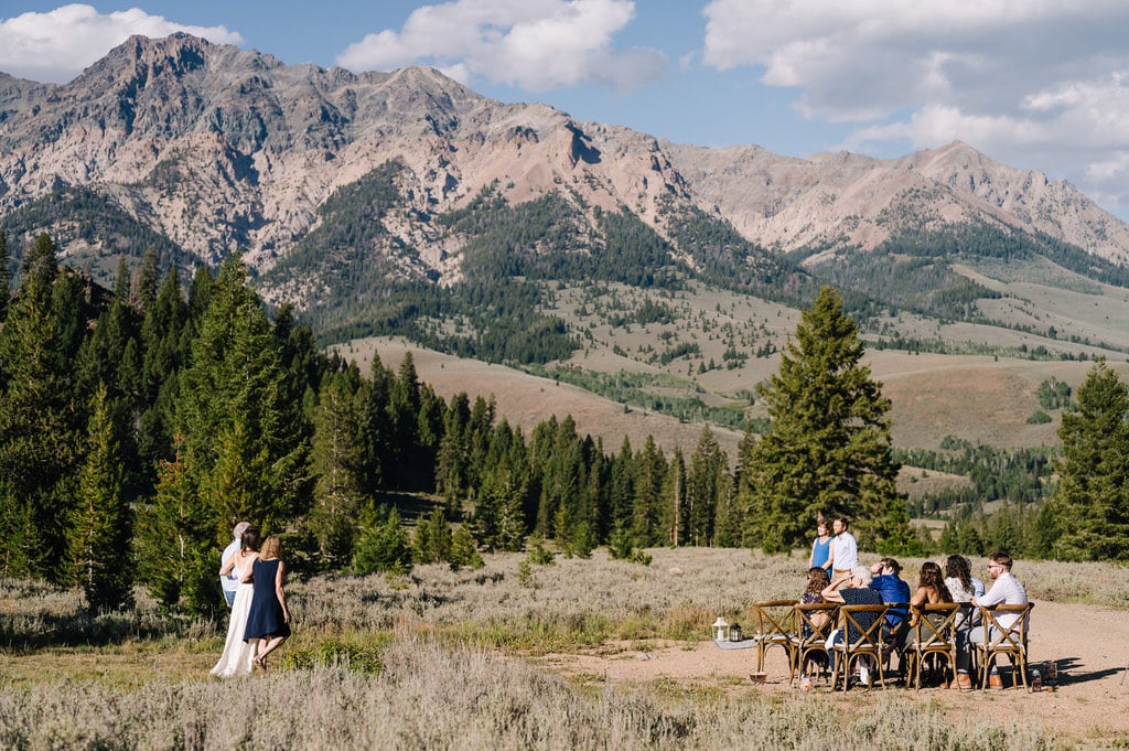 Mother and father walk their daughter down the aisle at her Sun Valley elopement. They are walking arm-in-arm and across a clearing and there are mountains behind them. Family and friends are seated to the side of the photo and the groom is standing in front.  
