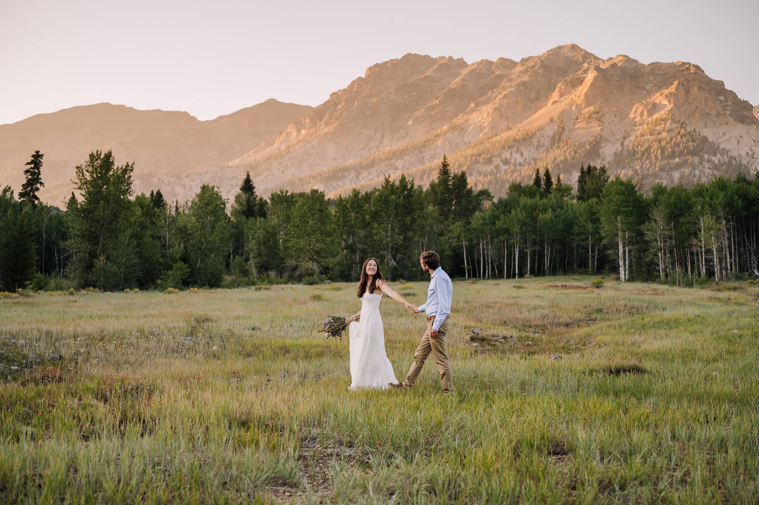 Couple walks across a meadow at sunset during their Sun Valley elopement. Bride is holding her groom's hand and looking back at him smiling. The setting sun is casting an alpenglow on the mountains behind the couple.