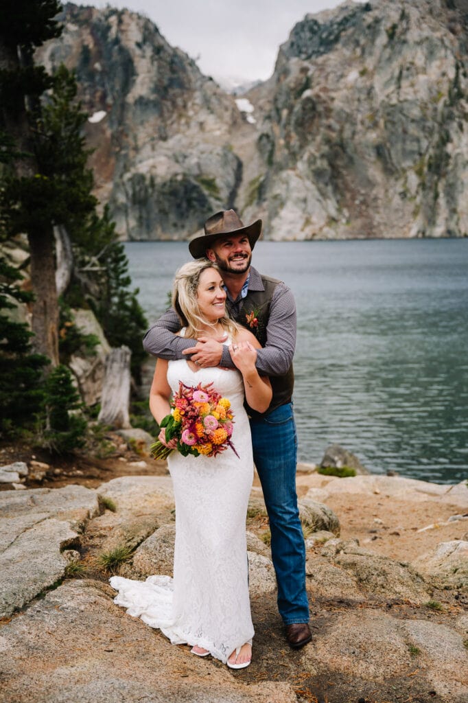 A couple stands by an alpine lake during their Idaho elopement package with Autumn Lynne Photography. Bride is wearing a white dress and holding a bouquet with orange flowers. Groom is wearing blue jeans, a vest and leather hat.