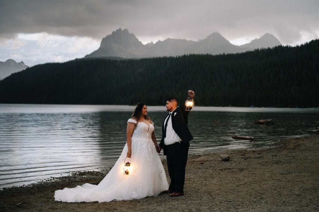 A couple stands together holding lanterns at dusk. The mountains behind them are covered by mist. This couple eloped in Stanley at Redfish Lake.