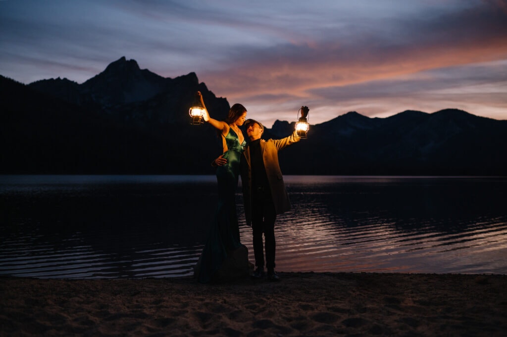 Autumn, an Idaho elopement photographer, captures a couple standing with lanterns on a mountain lake in Stanley. The sunset is illuminating the sky and the lake behind the couple.