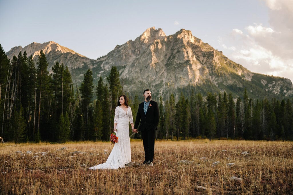 Autumn, an Idaho elopement photographer, captures a couple standing in an alpine meadow in Stanley, Idaho. The setting sun is highlighting the mountain behind the couple and they are facing the camera.
