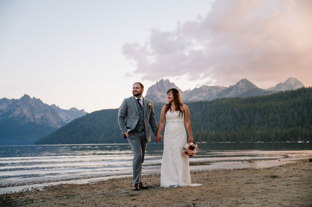 A wedding couple stands on a beach in Idaho with an alpine lake and rugged mountains behind them. The groom is wearing a grey suit and the bride is wearing a white wedding dress with a small train. Idaho is one of the easiest states to get married in. 
